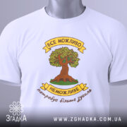 Футболка сила можливостей. Біла футболка, складена на плоскій поверхні, з видимим етикеткою. Принт з деревом та написом розташований по центру, фон світло-сірий, а футболка має округлий виріз.