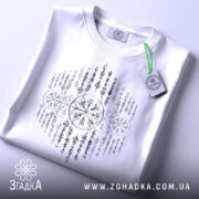 Футболка сила предків. Біла футболка, складена на поверхні, з видимим принтом у чорному кольорі. На футболці видно етикетку з зеленим шнурком, що додає акценту. Фон світло-сірий, що створює контраст з футболкою.