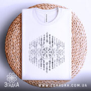 Футболка сила предків. Біла футболка, складена на плетеній основі, з видимим принтом у чорному кольорі. Фон світло-сірий, що створює м'який контраст з футболкою.