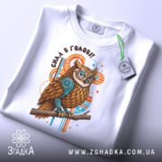 Футболка сила в голові, біла, зображена зверху, на ній прикріплений етикет, фон світлий, принт з совою з коричневим пір'ям та блакитними елементами, текст 'Сила в голові!!' над совою.