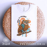 Футболка сила в голові, біла, зображена зверху на плетеній основі, фон світлий, принт з совою з коричневим пір'ям та блакитними елементами, текст 'Сила в голові!!' над совою.