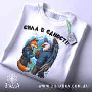 Футболка сила в єдності, біла, зображена зверху, фон світло-сірий, принт з лисицею та орлом, які стискають руки, на лисиці броня, на орлі светр, на футболці є етикетка.