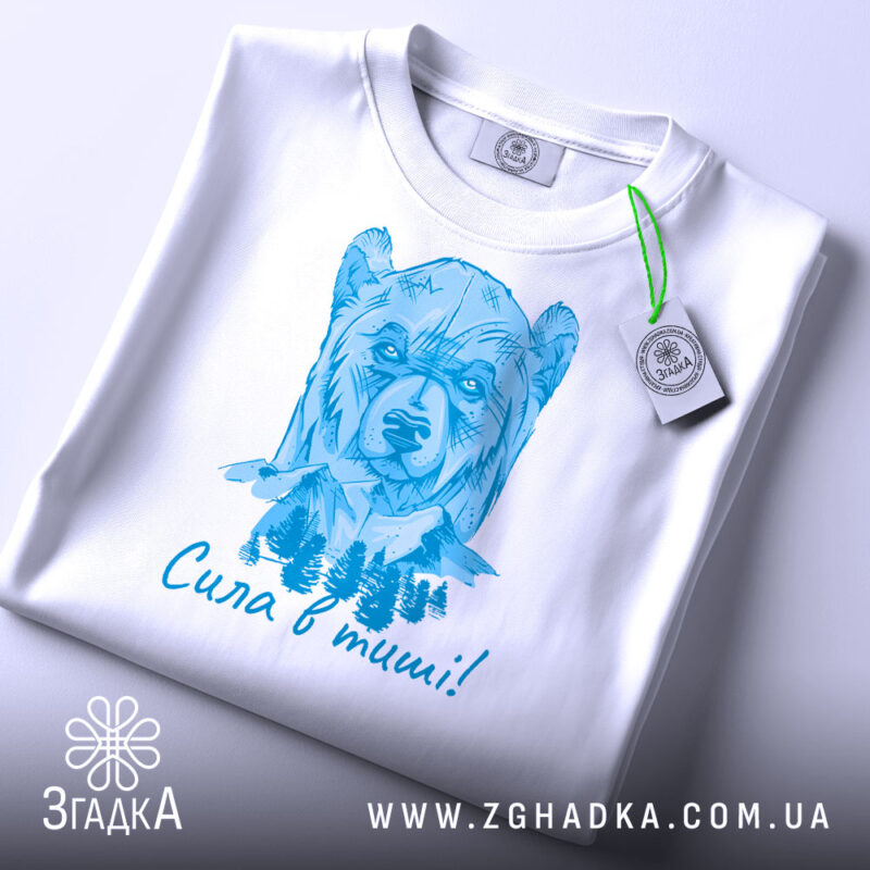 Футболка сила в тиші. Біла футболка, складена на поверхні, з малюнком ведмедя в блакитних тонах. На футболці видно етикетку з зеленим шнурком. Фон світлий, що створює чистий вигляд.