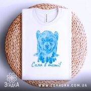 Футболка сила в тиші. Біла футболка, складена на плетеній кошику, з малюнком ведмедя в блакитних тонах. Фон світлий, що додає елегантності.