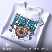 Футболка сила вікінга, біла, складена на столі, фон світло-сірий, принт з написом 'VIKING' у зелених та чорних кольорах, з зображенням щита та сокири, з етикеткою на зеленій стрічці.