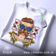 Футболка сила волі. Складена біла футболка з принтом черепа в окулярах, на фоні вишиванки. Вид з верхнього ракурсу, з етикеткою на комірці. Принт кольоровий, з елементами рослинності.