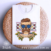 Футболка сила волі. Складена біла футболка з принтом черепа, що носить окуляри, на фоні плетеного кошика. Вид зверху, принт кольоровий, з елементами рослинності та написом 'GLORY TO UKRAINE'.