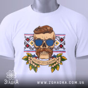 Футболка сила волі. Складена біла футболка з принтом черепа в окулярах, на фоні вишиванки. Вид з верхнього ракурсу, з етикеткою на комірці. Принт кольоровий, з елементами рослинності.