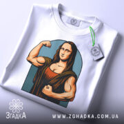 Футболка сильна моналіза, біла, складена на столі, фон світло-сірий, принт зображає Моналізу з м'язами, що позує з кулаком, з етикеткою зеленого кольору.