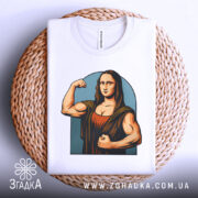 Футболка сильна моналіза, біла, складена на плетеній кошику, фон світло-сірий, принт зображає Моналізу з м'язами, що позує з кулаком, в кольорах коричневого та блакитного.