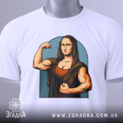 Футболка сильна моналіза, біла, складена на столі, фон світло-сірий, принт зображає Моналізу з м'язами, що позує з кулаком, з етикеткою зеленого кольору.