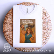 Футболка сильніша ніж думаю, біла, складена на плетеному кошику, фон світло-сірий, принт з жінкою та чоловіком, з написом 'СИЛЬНІША, НІЖ ДУМАЮ', видно етикетку на комірці.