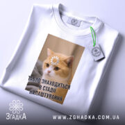 Футболка табло налаштовується, біла, зображена з верхнього ракурсу, фон світлий, принт з котом, на якому квітка на лобі та текст "ТАБЛЮ ЗНАХОДИТЬСЯ В СТАДІ НАЛАШТУВАННЯ", на комірці є етикетка.