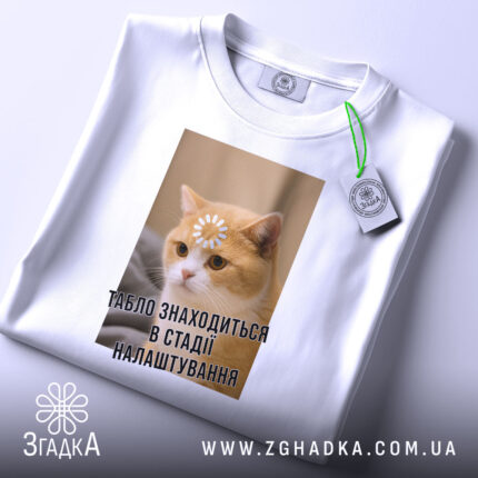 Футболка табло налаштовується, біла, зображена з верхнього ракурсу, фон світлий, принт з котом, на якому квітка на лобі та текст "ТАБЛЮ ЗНАХОДИТЬСЯ В СТАДІ НАЛАШТУВАННЯ", на комірці є етикетка.