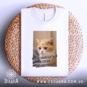 Футболка табло налаштовується, біла, зображена з верхнього ракурсу, фон плетений, принт з котом, на якому квітка на лобі та текст "ТАБЛЮ ЗНАХОДИТЬСЯ В СТАДІ НАЛАШТУВАННЯ".