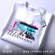 Футболка Тарас Григорович Шевченко, біла, складена на поверхні, фон світло-сірий, принт з портретом Шевченка, кольорові акценти в блакитному та рожевому, на комірці є етикетка.