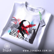 Футболка Тарас Шевченко білого кольору, зображена на столі, ракурс зверху, фон світлий, принт з портретом Тараса Шевченка, текстовий елемент та яскраві кольорові акценти, на футболці є бирка.