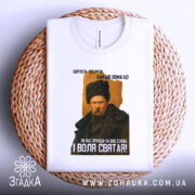 Футболка Тарас Шевченко борітеся, біла футболка, складена на плетеній основі, зображення Тараса Шевченка в чорній шапці, фон білий, текст на футболці чорний і жовтий.