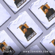 Футболка Тарас Шевченко борітеся, біла футболка з принтом, зображення Тараса Шевченка в чорній шапці, фон світло-сірий, текст на футболці чорний і жовтий, розташування принта по центру.