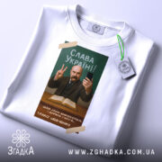 Футболка Тарас Шевченко селфі, біла, зображена зверху, фон світлий, принт з портретом Тараса Шевченка, він тримає телефон і показує знак миру, текст "Слава Україні!" на зеленому фоні, з етикеткою.