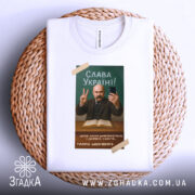 Футболка Тарас Шевченко селфі, біла, зображена зверху на плетеній основі, фон світлий, принт з портретом Тараса Шевченка, він тримає телефон і показує знак миру, текст "Слава Україні!" на зеленому фоні.