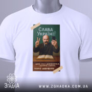 Футболка Тарас Шевченко селфі, біла, зображена зверху, фон світлий, принт з портретом Тараса Шевченка, він тримає телефон і показує знак миру, текст "Слава Україні!" на зеленому фоні, з етикеткою.