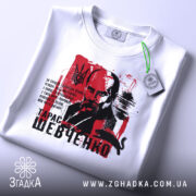 Футболка Тарас Шевченко білого кольору, зображена зверху, складена на плоскій поверхні, фон світлий, принт з портретом Шевченка на червоному фоні, з зеленим ярликом.