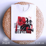 Футболка Тарас Шевченко білого кольору, зображена зверху, складена на плоскій поверхні з плетеним елементом, фон світлий, принт з портретом Шевченка на червоному фоні.