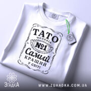 Футболка тато чоловік білого кольору, зображена зверху, на плоскій поверхні, фон світло-сірий, принт чорного кольору з написом 'TATO чоловік №1 Самий кращий у світі', на футболці є етикетка з зеленим шнурком.
