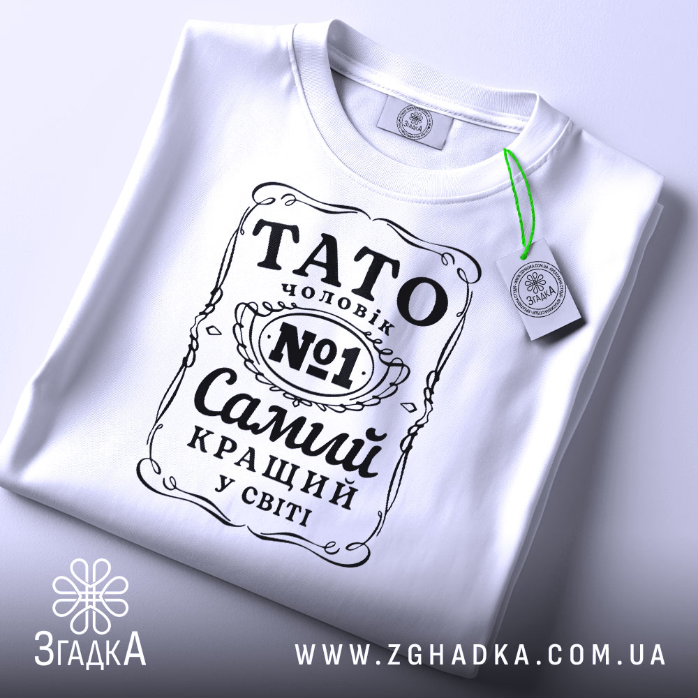 Футболка тато чоловік білого кольору, зображена зверху, на плоскій поверхні, фон світло-сірий, принт чорного кольору з написом 'TATO чоловік №1 Самий кращий у світі', на футболці є етикетка з зеленим шнурком.