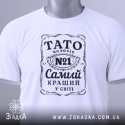 Футболка тато чоловік білого кольору, зображена зверху, на плоскій поверхні, фон світло-сірий, принт чорного кольору з написом 'TATO чоловік №1 Самий кращий у світі', на футболці є етикетка з зеленим шнурком.