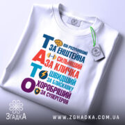 Футболка тато герой для дітей, біла, зображена зверху, на столі, фон світло-сірий, принт кольоровий з текстом "ТАТО" та іконками, на футболці є етикетка з логотипом.