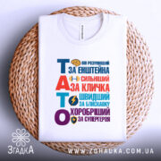 Футболка тато герой для дітей, біла, зображена зверху на плетеній кошику, фон світло-сірий, принт кольоровий з текстом "ТАТО" та іконками, що підкреслюють його значення.