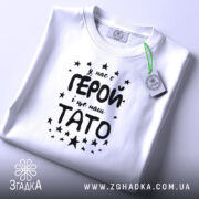 Футболка тато герой, біла, складена на поверхні, фон світло-сірий, принт чорного кольору з текстом 'У нас є ГЕРОЙ і це наш ТАТО' оточений зірочками, на футболці є етикетка з зеленим шнурком.