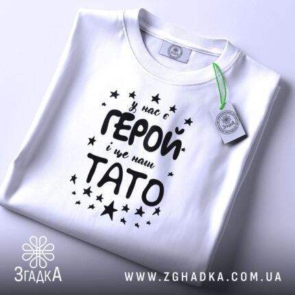 Футболка тато герой, біла, складена на поверхні, фон світло-сірий, принт чорного кольору з текстом 'У нас є ГЕРОЙ і це наш ТАТО' оточений зірочками, на футболці є етикетка з зеленим шнурком.