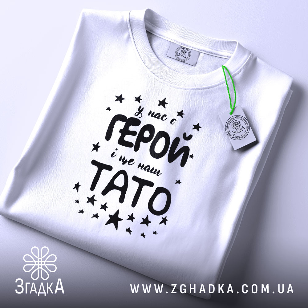 Футболка тато герой, біла, складена на поверхні, фон світло-сірий, принт чорного кольору з текстом 'У нас є ГЕРОЙ і це наш ТАТО' оточений зірочками, на футболці є етикетка з зеленим шнурком.