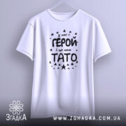 Футболка тато герой, біла, підвішена на вішалці, фон світло-сірий, принт чорного кольору з текстом 'У нас є ГЕРОЙ і це наш ТАТО' оточений зірочками.
