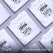 Футболка тато герой, біла, зображена на манекені, фон світло-сірий, принт чорного кольору з текстом 'У нас є ГЕРОЙ і це наш ТАТО' оточений зірочками.