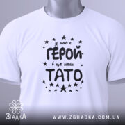 Футболка тато герой, біла, складена на поверхні, фон світло-сірий, принт чорного кольору з текстом 'У нас є ГЕРОЙ і це наш ТАТО' оточений зірочками, на футболці є етикетка з зеленим шнурком.