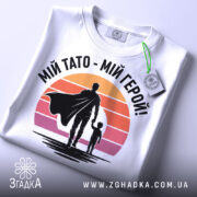 Футболка тато герой, біла, складена на поверхні, фон світло-сірий, принт з фігурами батька та дитини на фоні градієнту від оранжевого до рожевого, з етикеткою на рукаві.