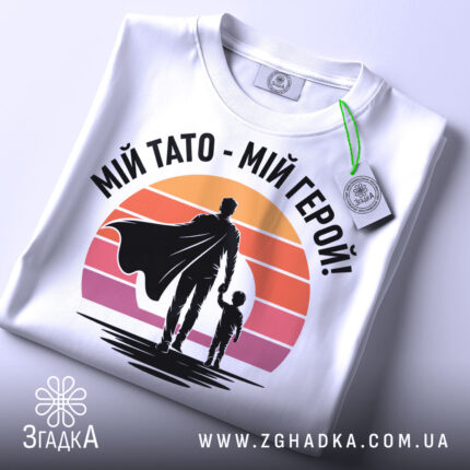 Футболка тато герой, біла, складена на поверхні, фон світло-сірий, принт з фігурами батька та дитини на фоні градієнту від оранжевого до рожевого, з етикеткою на рукаві.