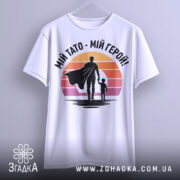 Футболка тато герой, біла, підвішена на вішалці, фон світло-сірий, принт з фігурами батька та дитини на фоні градієнту від оранжевого до рожевого.