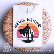 Футболка тато герой, біла, складена на плетеній кошику, фон світло-сірий, принт з фігурами батька та дитини на фоні градієнту від оранжевого до рожевого.