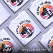 Футболка тато герой, біла, зображена на манекені, фон світло-сірий, принт з фігурами батька та дитини на фоні градієнту від оранжевого до рожевого.