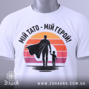 Футболка тато герой, біла, складена на поверхні, фон світло-сірий, принт з фігурами батька та дитини на фоні градієнту від оранжевого до рожевого, з етикеткою на рукаві.