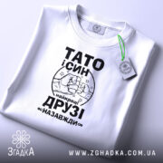 Футболка тато і син білого кольору, складена на поверхні, фон світло-сірий, принт чорного кольору з написом 'ТАТО і СИН найкращі ДРУЗІ назавжди' та малюнком рук, що стискаються, з зеленим ярликом.