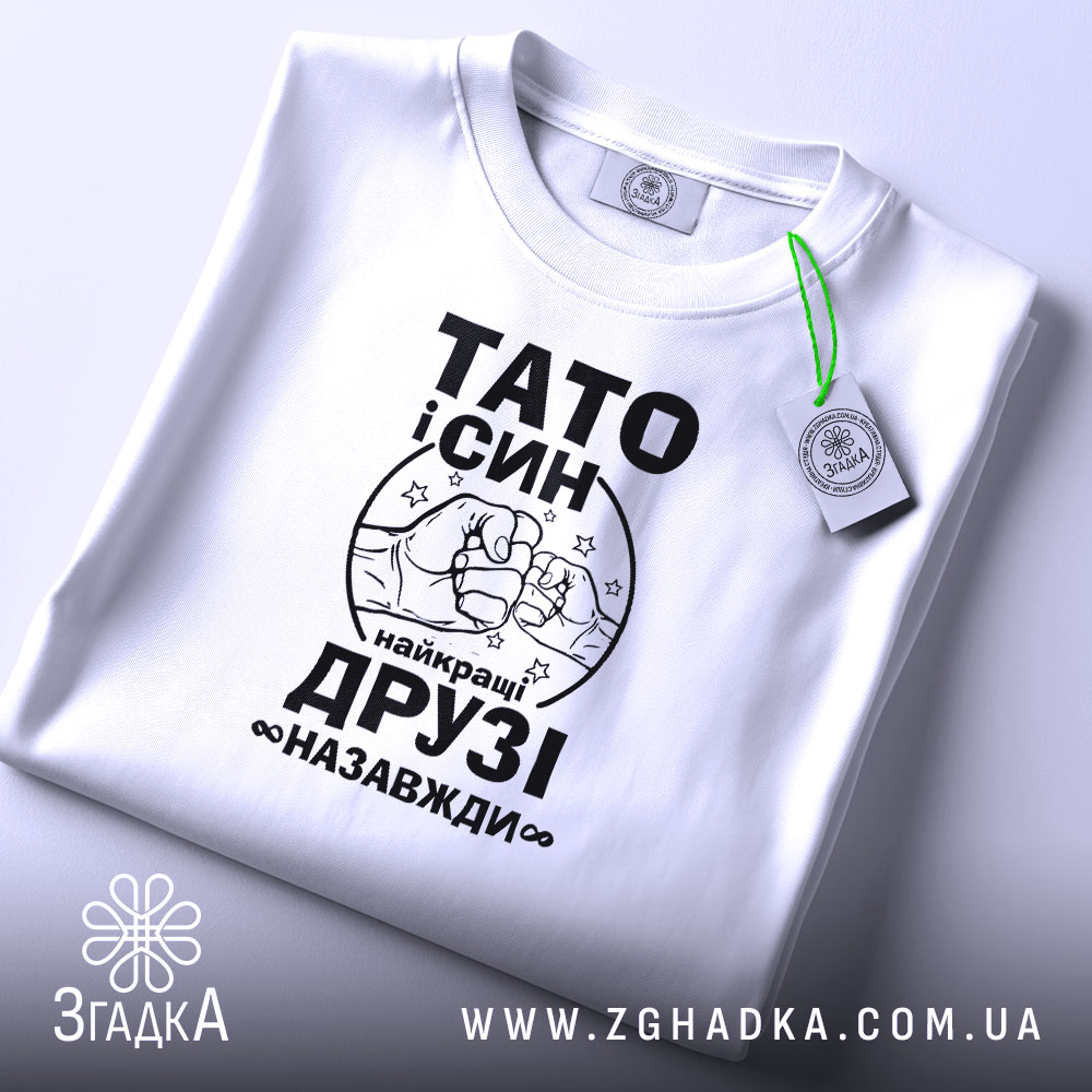 Футболка тато і син білого кольору, складена на поверхні, фон світло-сірий, принт чорного кольору з написом 'ТАТО і СИН найкращі ДРУЗІ назавжди' та малюнком рук, що стискаються, з зеленим ярликом.