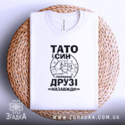 Футболка тато і син білого кольору, складена на плетеній кошику, фон білий, принт чорного кольору з написом 'ТАТО і СИН найкращі ДРУЗІ назавжди' та малюнком рук, що стискаються.