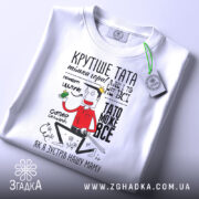 Футболка тато може все, біла, лежить на столі, фон світлий, принт з персонажем, що тримає зелений предмет, текст на футболці яскравий і кумедний.