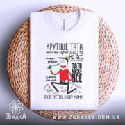 Футболка тато може все, біла, лежить на плетеному кошику, фон нейтральний, принт з персонажем, що тримає зелений предмет, текст на футболці яскравий і привітний.